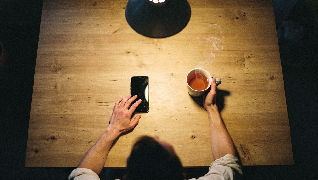 A person sitting at a kitchen table with their phone face-up beside them, hand resting near it but not picking it up, soft evening light